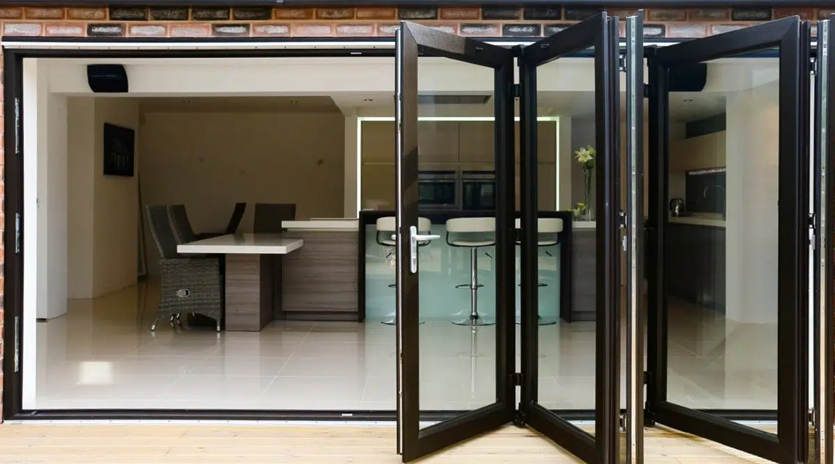 A dark-framed WeatherMaster™ Plus Bi-Fold Door system partially opened, connecting a modern indoor kitchen and dining area to an outdoor wooden deck.